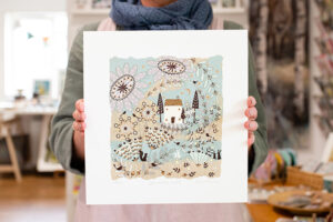 Picture of a staff member holding a square art print featuring a illustrated house with floral elements around it.
