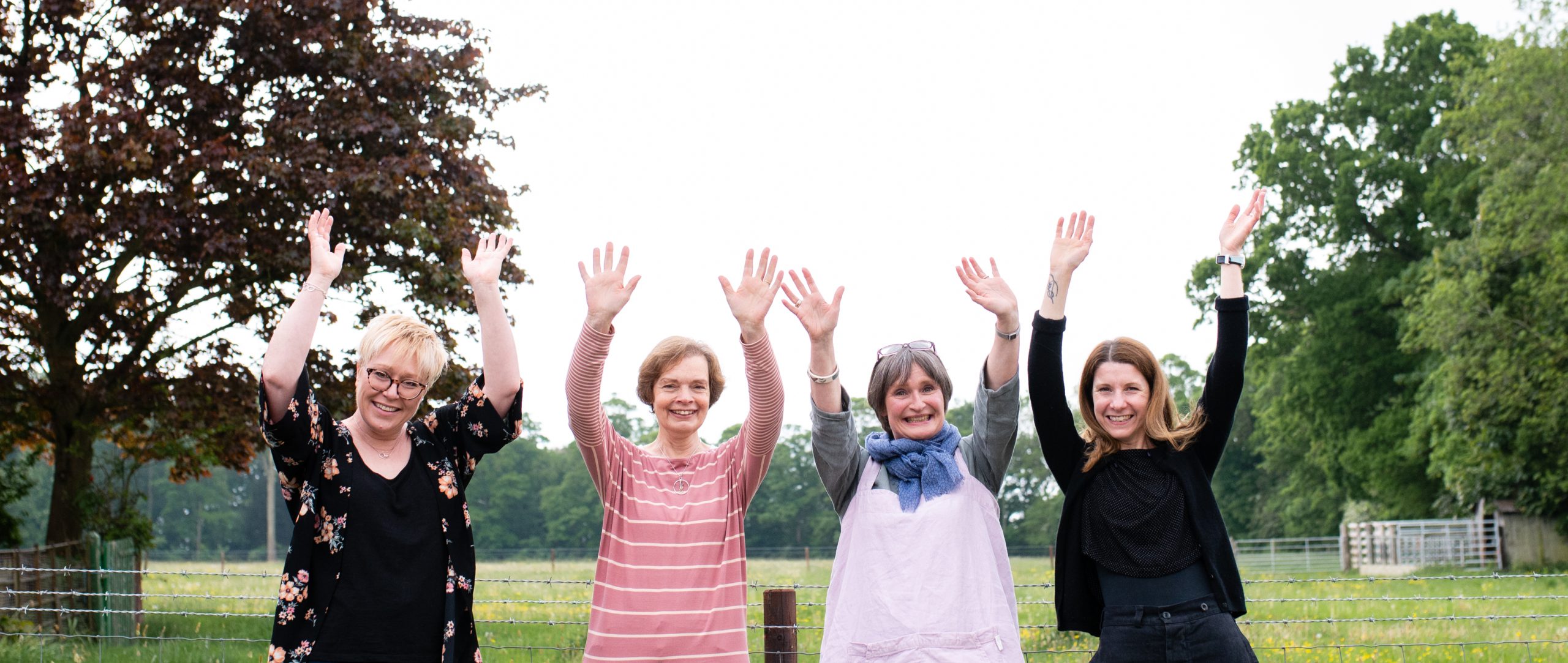 Members of wistow gallery outside waving there hands up in the air with big smiles on their faces and wistow countryside behind them.