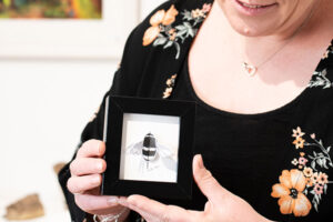 Picture of staff member holding small black frame with a 3d paper made bee inside.