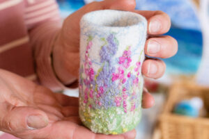 Picture of felt/textured handmade small vase/pot featuring a wildflower plants with lilacs pinks, greens and whites. Very fluffy in look and textured.