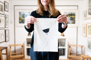 Sarah gallery owner hold fabric tapestry featuring a small bird made out of thread.