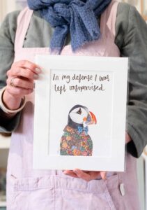 Picture of puffin art print with coloured suit on and and blue bow tie all hand illustrated in a white card frame. Above the puffin in hand illustrated calligraphy it reads in my defense I was left unsupervised.