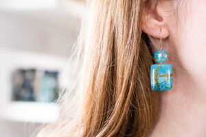 Picture of ear with a handmade blue earing with a circular first element and a rectangular dangling second element.