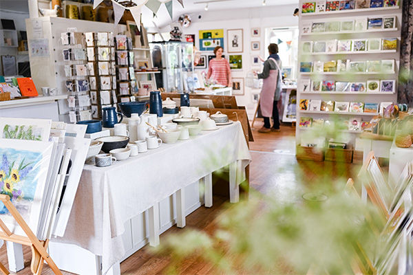 Picture of the inside of the gallery showcase lots of products from art prints, ceramics to greeting cards. In the far background a staff member is picture talking to a member of the public.