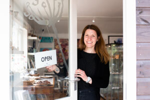 Picture of sarah the owner of wistow gallery partially holding the open sign on the entrance door with a beaming smile welcoming you to the gallery.