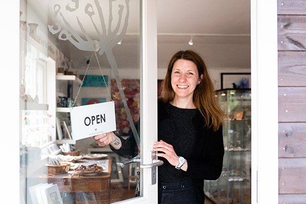 Picture of sarah the owner of wistow gallery partially holding the open sign on the entrance door with a beaming smile welcoming you to the gallery.