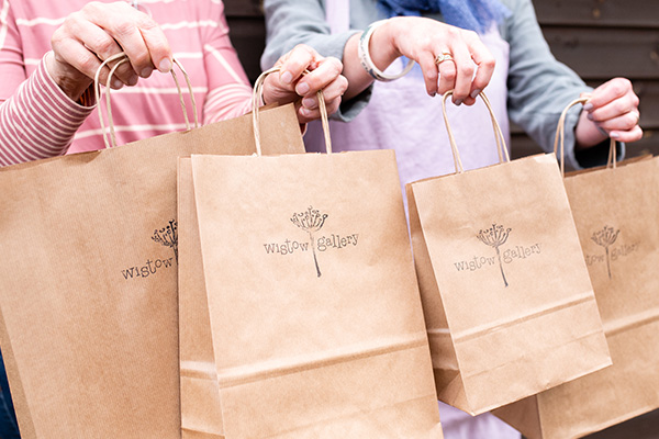 Picture of two wistow gallery staff members holding different sized kraft sustainable bags with wistow galleries logo stamped on them to ensure the environmental impact is reduced.