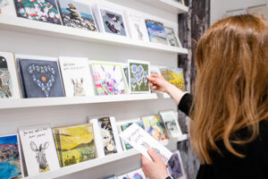 Picture of a person picking greeting cards from a display unit on the wall featuring an array of hand illustrated printed gift cards of varying sizes.