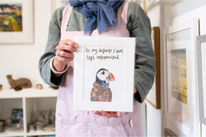 Art print in a white frame featuring a puffin with a floral coat which is hand illustrated. Text above the puffin in a calligraphy font reads in my defence I was left unsupervised
